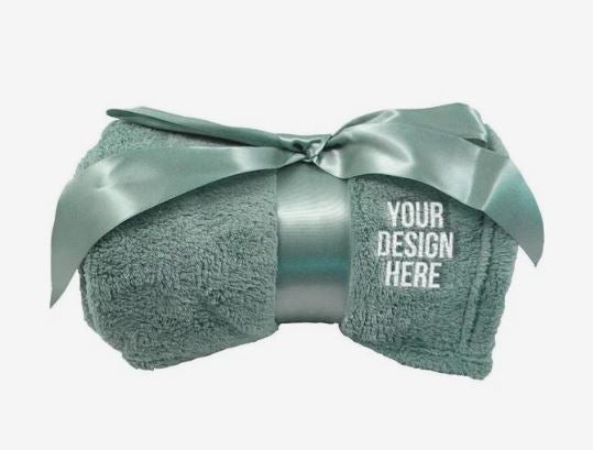 A photo of a plush sage green blanket rolled up and tied with a satin bow. YOUR DESIGN HERE is embroidered in the corner.
