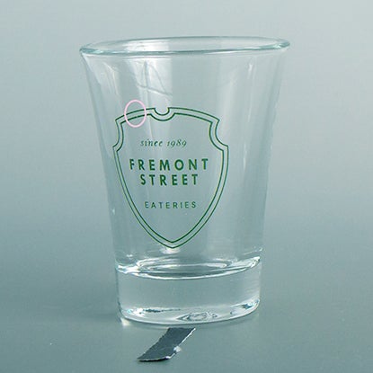 A photo of a shot glass with a green shield print design, with a small chip in the shield.