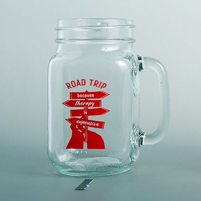A photo showing a glass mason jar with a handle. The jar is printed with a red road and road sign, and ROAD TRIP in red.