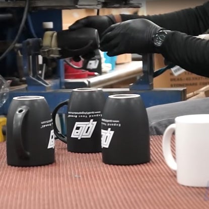 A photo showing a person wearing black latex gloves using a machine to screen print the QLP logo and company information in white on a black mug.