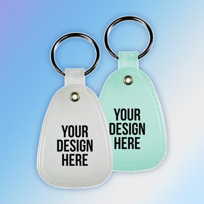 A photo of saddle-style faux leather glow-in-the-dark key tags, one purple-grey and one blue, both printed with YOUR DESIGN HERE. They're on a blue background.