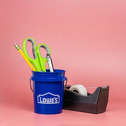 A photo showing a blue mini Lowe’s bucket filled with scissors, a pencil, and a pen, next to a sticky tape dispenser, on a pink background.
