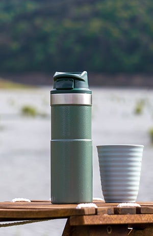 A photo showing a green flask next to a white ceramic cup. They are sitting on a wooden table with a lake and forest in the background.