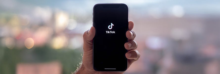 A photo of a hand holding a smartphone with a black case, with the screen showing the TikTok logo on a black background. In the background behind the hand is an out of focus cityscape.