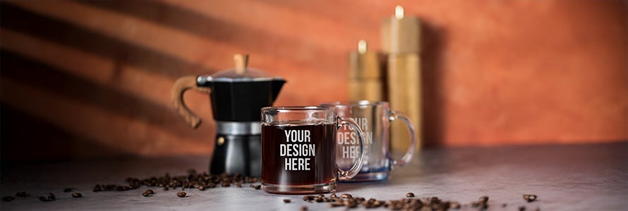 A photo showing two glass mugs printed with YOUR DESIGN HERE on the front, resting on a grey surface with a black metal moka pot in the background.