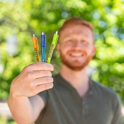 A photo of a smiling man holding three pens, in blue, orange, and green towards the camera.