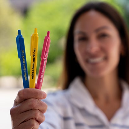 A photo of a smiling woman holding three pens, in blue, yellow, and pink towards the camera.