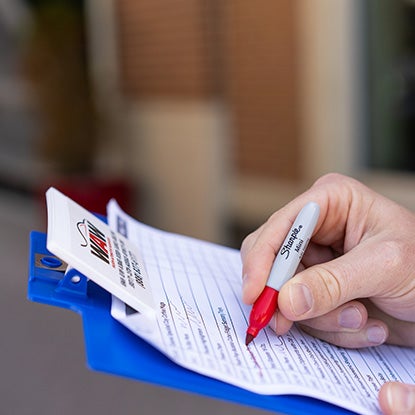 A photo showing someone using a mini red Sharpie to write on a piece of paper clipped to a blue clipboard.