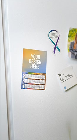 A photo showing a  white fridge with magnets stuck to it, including a rectangular magnet printed with a calendar.