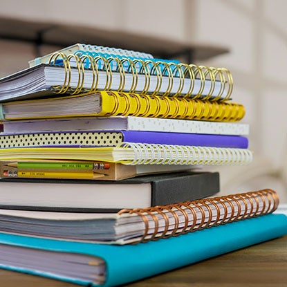 A photo of a pile of notebooks on a wooden desk, They’re a mix of case bound and wire-o bindings, all in different sizes and colors.