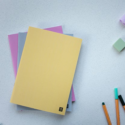 A photo showing a stack of three slim saddle stitch notebooks, with yellow, blue, and pink covers respectively. Each has a small black square logo in the bottom right corner of the cover. They are resting on a white surface, near some pens and erasers.
